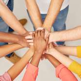 A group of diverse people stand in a circle with their hands stacked together in the center, symbolizing unity and teamwork.