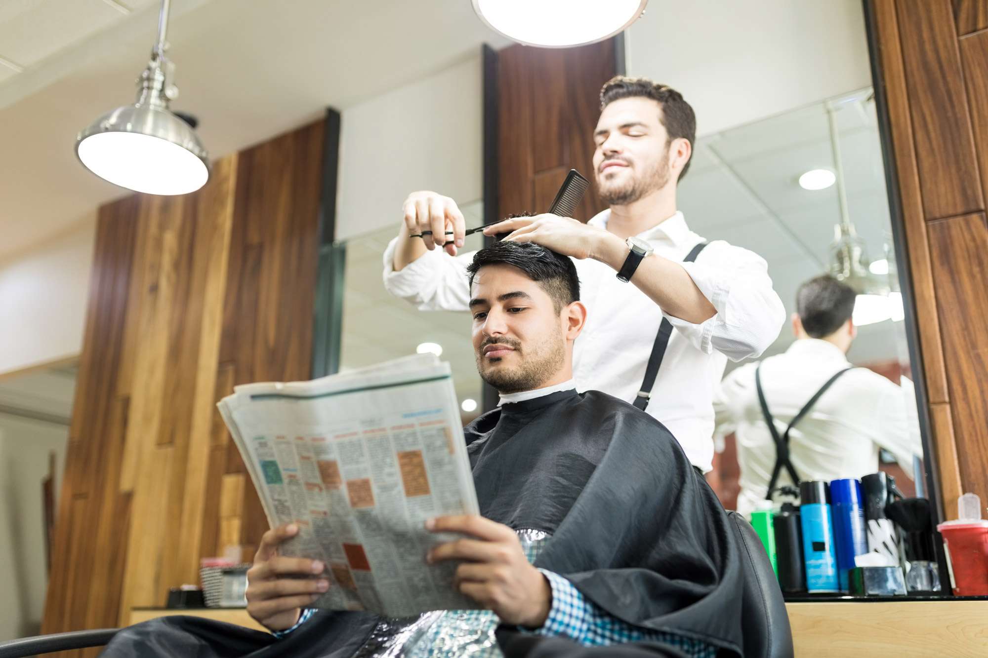 A man sits in a barber chair reading a newspaper while a barber stands behind him, cutting his hair. The salon has wooden panels, mirrors, and hair products on the counter.